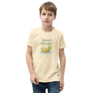 Può includere: T-shirt beige chiaro con la scritta "Speed Reader" in verde, sopra una tartaruga dei cartoni animati che legge un libro. La tartaruga è in una vasca di legno che galleggia sull'acqua blu. La maglietta è indossata da una persona giovane.