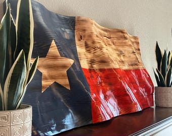 May include: A wooden Texas flag with a waving design. The flag features a large tan star on a blue background, with red and tan stripes. The wood grain is visible, adding texture. The flag is displayed on a shelf with potted plants.
