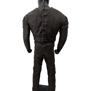 May include: A brown jumpsuit with white stitching and diamond-patterned accents on the sleeves. The suit has a high collar and is displayed on a mannequin. The back view shows the full garment.