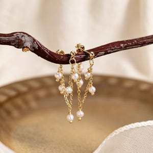 May include: Gold-colored dangle earrings with small, white pearls. The earrings are suspended from a brown branch against a blurred background. The earrings have a delicate chain design, with multiple pearls hanging at different lengths.