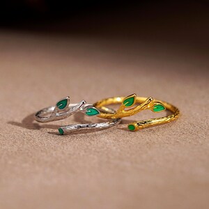May include: Two open-style rings shaped like tree branches, one in silver-tone metal and one in gold-tone metal. Each ring features textured bark detailing and small, bright green enamel leaves attached to the branch-like band structure.