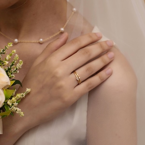 Könnte beinhalten: Nahaufnahme eines goldenen Rings mit einem kleinen Diamanten am Finger einer Person. Der Ring wird mit einer goldenen Perlenkette getragen. Ein Blumenstrauß ist in der linken unteren Ecke zu sehen. Die Person trägt einen weißen Schleier.
