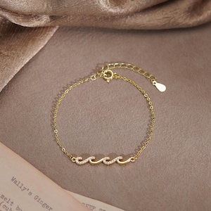 Puede incluir: Una pulsera de cadena en tono dorado con un dije central en forma de ola adornado con pequeñas piedras claras y brillantes. La pulsera incluye una cadena de eslabones finos, un cierre de resorte circular y una cadena extensora ajustable con una pequeña etiqueta metálica en forma de gota al final.