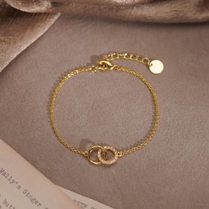 Könnte beinhalten: Goldfarbenes Armband mit zwei ineinandergreifenden Kreisen, von denen einer mit kleinen, klaren Kristallen besetzt ist. Die feine Kette hat eine verstellbare Länge und einen kleinen, runden Anhänger. Das Schmuckstück liegt auf einer neutralen Oberfläche.