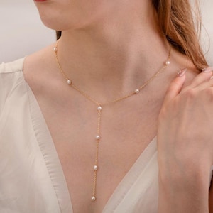 Puede incluir: Un delicado collar de oro con pequeñas perlas redondas. El collar tiene un diseño en forma de Y, con perlas espaciadas a lo largo de la cadena y una hebra más larga de perlas colgando. El collar se usa con un vestido blanco.