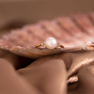 May include: A gold ring with a large, white pearl and small accent stones. The ring is set on a seashell, against a soft, neutral background. The pearl is the focal point of the ring, which is a classic piece of jewellery.