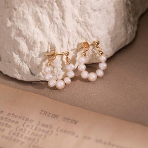 Può includere: Un paio di orecchini a cerchio dorati adornati con piccole perle lucenti. Gli orecchini sono esposti su uno sfondo strutturato bianco sporco, con un libro di ricette vintage visibile. Gli orecchini hanno un design classico ed elegante.