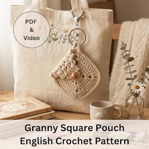 Puede incluir: Una bolsa cuadrada de ganchillo beige con cierre de botón cuelga de un llavero plateado. La bolsa está sujeta a un bolso tote beige con bordados florales. El texto "PDF & Video" está en un círculo en la bolsa. Las palabras "Granny Square Pouch English Crochet Pattern" están en la parte inferior.