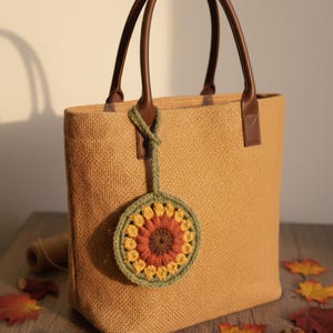 Könnte beinhalten: Eine beigefarbene Tragetasche mit braunen Ledergriffen und einem gehäkelten Sonnenblumen-Anhänger. Die Tasche hat eine strukturierte, gewebte Optik. Der Sonnenblumen-Anhänger hat gelbe, orange und braune Farben mit einem grünen Rand.