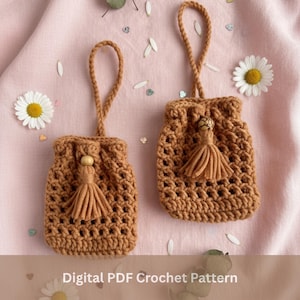 May include: Two brown crochet drawstring bags with tassels and wooden beads. Each bag has a loop handle. The bags are displayed on a pink surface with daisy flowers and petals. The text "Digital PDF Crochet Pattern" is at the bottom.