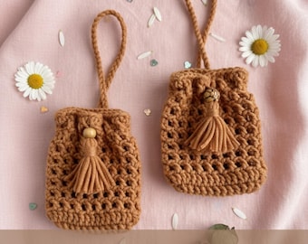 May include: Two brown crochet drawstring bags with tassels and wooden beads. Each bag has a loop handle. The bags are displayed on a pink surface with daisy flowers and petals. The text "Digital PDF Crochet Pattern" is at the bottom.