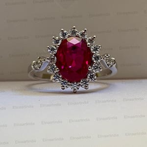 Op de afbeelding: Een zilverkleurige ring met een grote, ovaal geslepen, dieprode edelsteen in het midden. De centrale steen is omringd door een halo van kleine, ronde, helderwitte steentjes. Aan beide zijden van de ringband zijn extra heldere steentjes gezet.