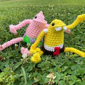 Puede incluir: Dos figuras de ganchillo hechas a mano de personajes de dibujos animados. Una es rosa con forma de estrella de mar, la otra es amarilla con forma cuadrada. Ambas figuras tienen ojos negros y están sentadas en hierba verde.