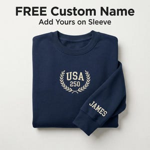 May include: A navy blue sweatshirt with the text "FREE Custom Name" and "Add Yours on Sleeve" at the top. The sweatshirt has "USA 250" embroidered on the chest and "JAMES" on the sleeve in a cream color.