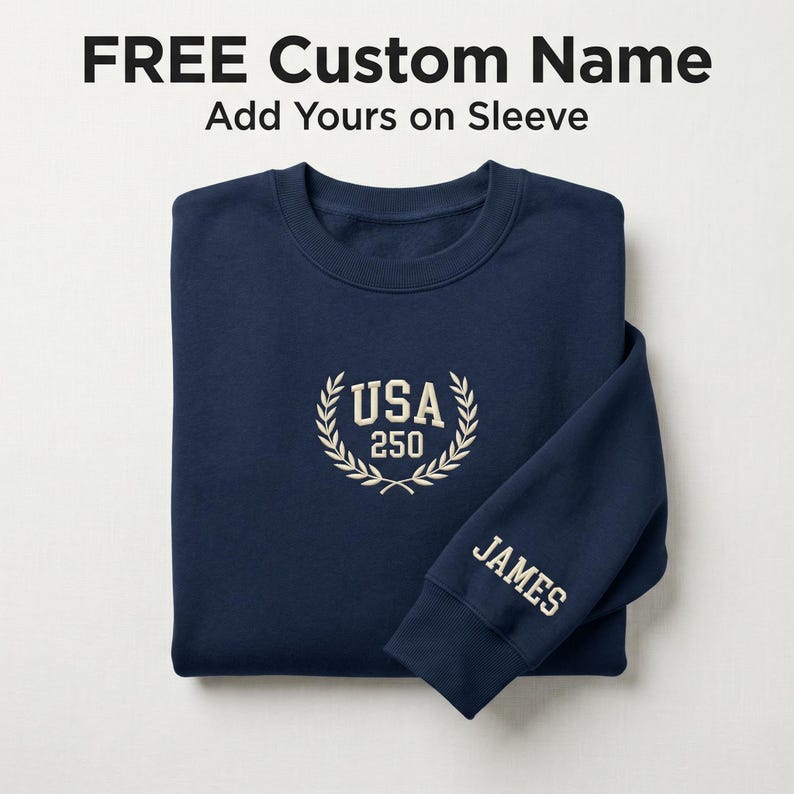 May include: A navy blue sweatshirt with the text "FREE Custom Name" and "Add Yours on Sleeve" at the top. The sweatshirt has "USA 250" embroidered on the chest and "JAMES" on the sleeve in a cream color.