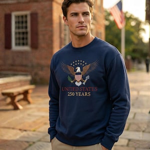 May include: Navy blue sweatshirt featuring an embroidered eagle design with the text "UNITED STATES 250 YEARS". The eagle is depicted with outstretched wings, surrounded by stars. The sweatshirt is a crew neck style.