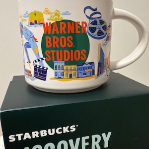 Puede incluir: Taza de cerámica blanca con el logotipo de Warner Bros. Studios y varias ilustraciones de dibujos animados. La taza está colocada sobre una caja verde oscuro de Starbucks Discovery Series. La taza tiene un asa curva.
