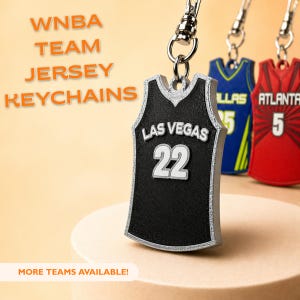 Puede incluir: Llavero de camiseta de Las Vegas negro y plateado con el número 22. El llavero forma parte de una colección de llaveros de camisetas de equipos de la WNBA. Otras camisetas de equipos son visibles en el fondo, incluyendo Dallas y Atlanta.