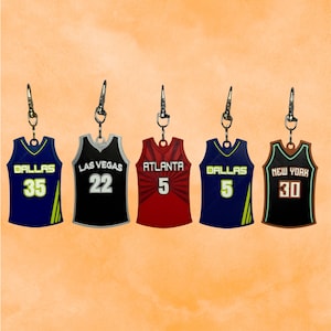 Puede incluir: Cinco llaveros de camisetas de baloncesto en azul marino, negro y rojo. Cada camiseta tiene un nombre y número de equipo diferente, incluyendo Dallas, Las Vegas, Atlanta y Nueva York. Los llaveros tienen cierres plateados.
