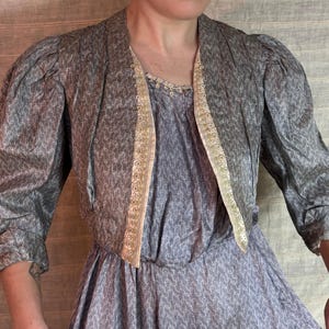 May include: A vintage-style dress and jacket set. The dress is a muted blue with a white pattern, and the jacket is a matching gray with three-quarter sleeves. The jacket is trimmed with a gold-colored lace.