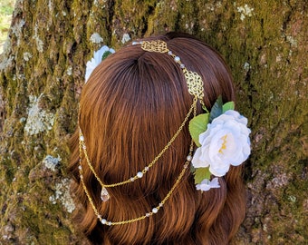 May include: A gold-colored headpiece with a filigree design and dangling chains, adorned with small pearls and a teardrop-shaped crystal. White fabric flowers with green leaves are attached to the side, creating a delicate, romantic look.