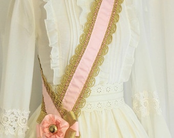 May include: A white dress with a pink and gold sash tied in a bow at the waist. The sash has a delicate lace trim.