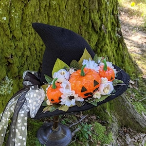 Puede incluir: Un sombrero de bruja negro de ala ancha decorado con flores blancas y detalles de calabaza naranja. El sombrero está sobre un soporte de madera.