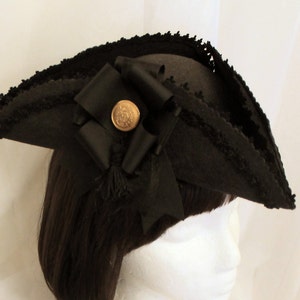May include: A black felt pirate hat with a black ribbon and a gold button. The hat has a fringed edge and is worn on a mannequin.