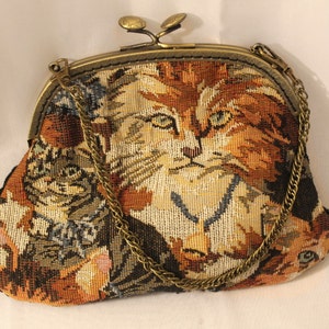 May include: A small tapestry purse with a gold chain strap. The purse features a cat face design with brown, orange, and white colours.