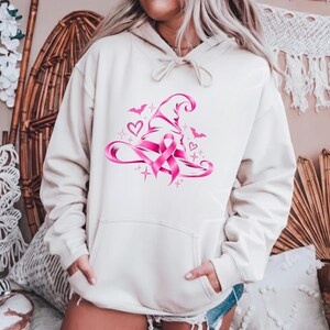 May include: A cream-coloured hooded sweatshirt featuring a graphic print of a pink witch hat intertwined with a breast cancer awareness ribbon. The design includes small pink hearts, stars, and bat silhouettes surrounding the central hat motif.