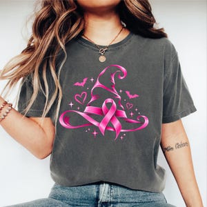 May include: A charcoal grey short-sleeve t-shirt featuring a vibrant pink graphic of a witch hat intertwined with a breast cancer awareness ribbon. The design includes small pink hearts, stars, and bat silhouettes surrounding the central hat motif.