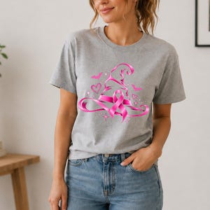 May include: A heather grey short-sleeved t-shirt featuring a bright pink graphic of a witch hat combined with a breast cancer awareness ribbon. The design includes small pink hearts, stars, and bat silhouettes surrounding the central hat motif.