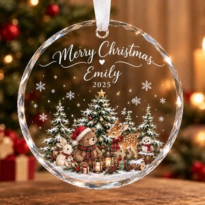 以下が含まれることがあります： Merry Christmas Emily 2025の文字が刻まれた、縁がカットされた円形の透明なガラス製オーナメント。テディベア、鹿、ウサギ、フクロウ、クリスマスツリーが描かれた森の風景がデザインされています。