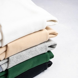 May include: A stack of five folded sweatshirts in solid colours. The top garment is white, followed by a beige sweatshirt, a heather grey one, a dark forest green piece, and a black sweatshirt at the bottom of the pile, all featuring a smooth texture.