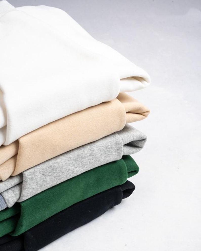 May include: A stack of five folded sweatshirts in solid colours. The top garment is white, followed by a beige sweatshirt, a heather grey one, a dark forest green piece, and a black sweatshirt at the bottom of the pile, all featuring a smooth texture.