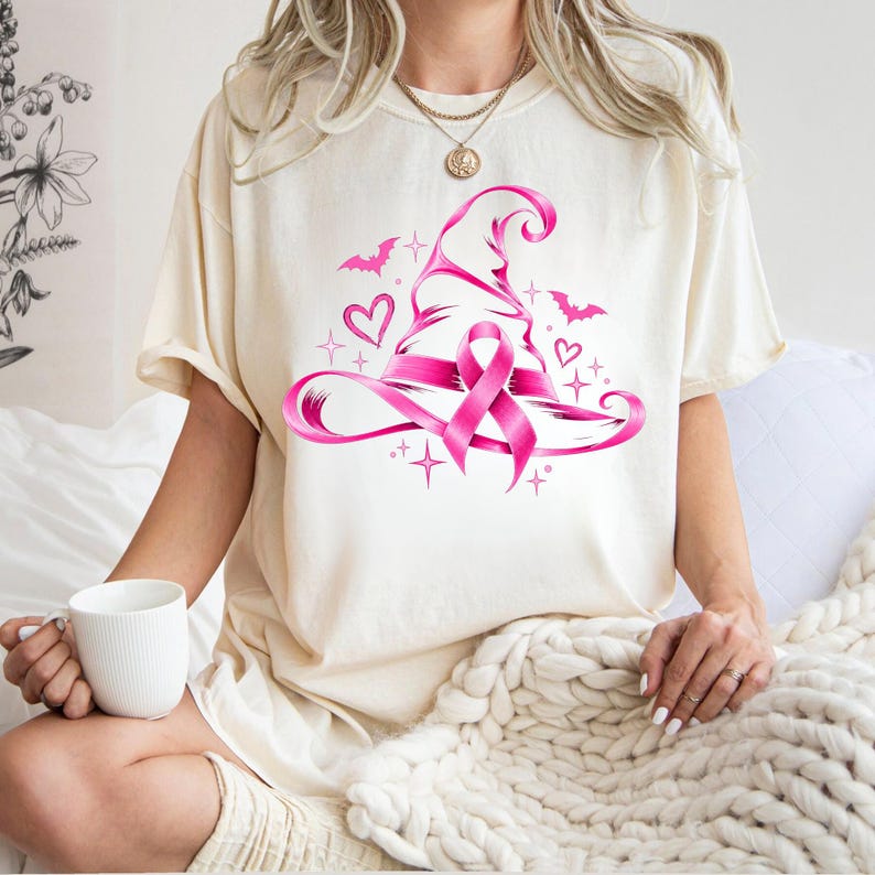 May include: A cream-coloured short-sleeve t-shirt featuring a graphic print of a bright pink witch hat adorned with a breast cancer awareness ribbon. The design includes small pink hearts, bats, and starburst accents surrounding the central hat.