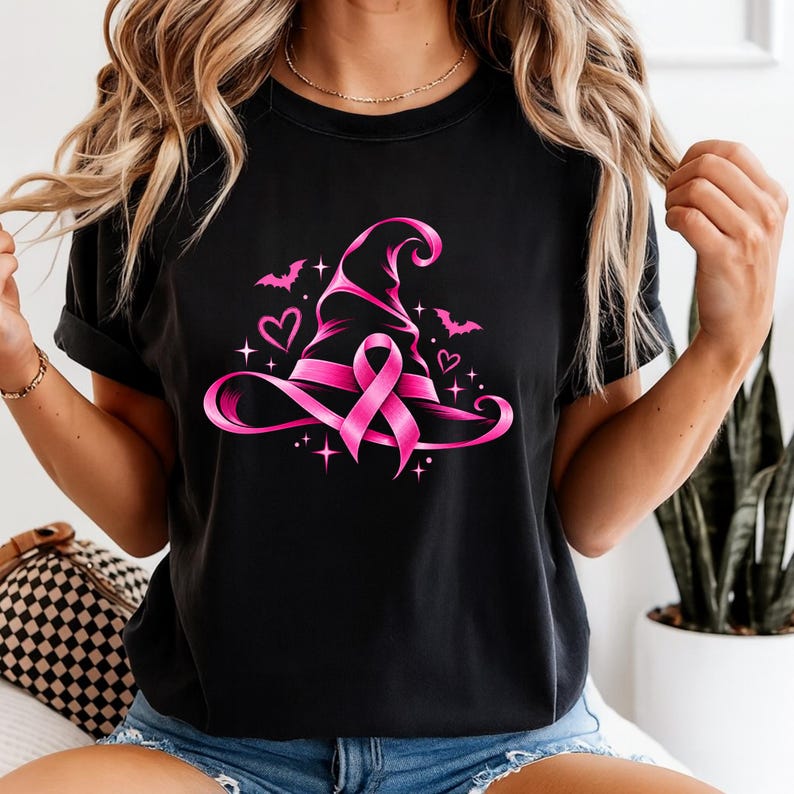 May include: A black short-sleeved t-shirt featuring a vibrant pink graphic of a witch hat adorned with a breast cancer awareness ribbon. The design includes small pink hearts, stars, and bat silhouettes surrounding the central hat illustration.