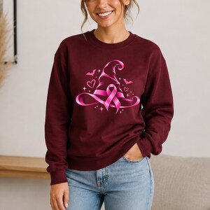 May include: A maroon long-sleeve sweatshirt featuring a central graphic of a bright pink witch hat intertwined with a breast cancer awareness ribbon. The design is accented with small pink hearts, flying bats, and sparkling star shapes.