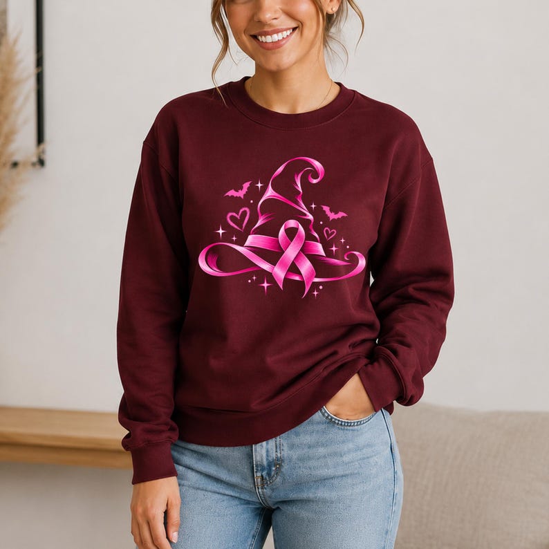May include: A maroon long-sleeve sweatshirt featuring a central graphic of a bright pink witch hat intertwined with a breast cancer awareness ribbon. The design is accented with small pink hearts, flying bats, and sparkling star shapes.