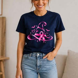 May include: A navy blue short-sleeved t-shirt featuring a graphic print of a pink witch hat formed by a breast cancer awareness ribbon. The design includes small pink hearts, stars, and bat silhouettes surrounding the central hat graphic.