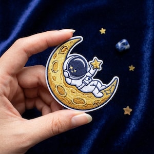 May include: Embroidered patch featuring an astronaut relaxing on a crescent moon. The astronaut is wearing a white spacesuit with a navy blue helmet. The moon is gold with embroidered details. The astronaut is reaching for a gold star. The patch is on a dark blue background.