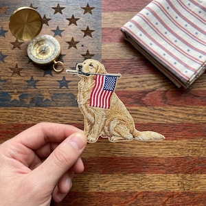 May include: Embroidered patch of a golden retriever dog holding a small American flag in its mouth. The dog is sitting, with detailed fur and a patriotic theme. The background features a wooden surface with an American flag design.