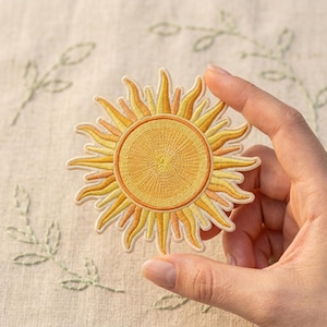 May include: Embroidered sun patch with a yellow and orange color scheme. The sun has a detailed, textured design with rays extending outward. The patch is likely for clothing or accessories, adding a decorative element.
