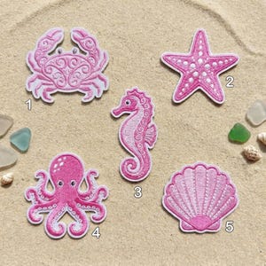 May include: A collection of five pink embroidered patches featuring sea creatures: a crab, starfish, seahorse, octopus, and seashell. Each patch has a white border and is set against a sandy background.