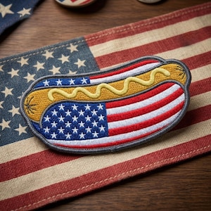 May include: Embroidered patch in the shape of a hot dog, featuring the American flag design. The hot dog has a yellow mustard topping. The patch is placed on a background of the American flag.