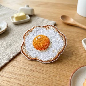 May include: Embroidered coaster in the shape of a fried egg. The coaster features a white center with an orange yolk and a brown, textured edge. The coaster is placed on a wooden surface.