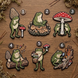 May include: A collection of six embroidered patches featuring whimsical green frogs in various poses. The frogs are depicted with mushrooms, musical instruments, and other forest-themed elements. The patches are on a wooden surface.