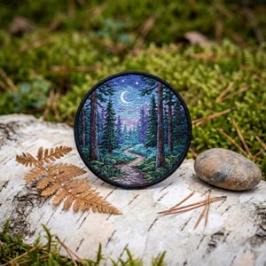 May include: Round embroidered patch featuring a forest scene with a path leading to the horizon under a starry night sky with a crescent moon. The patch has a black border and is set on a piece of birch bark.