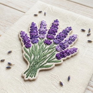 May include: Embroidered lavender bouquet patch featuring vibrant purple flower heads and green stems. The patch is on a cream-colored fabric, with loose lavender seeds scattered around. A charming floral accent for clothing or crafts.
