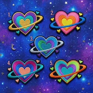 May include: Five embroidered heart patches with a planet-like design. Each heart features multiple layers of color, surrounded by a ring and small heart accents. The background is a starry, space-themed fabric.